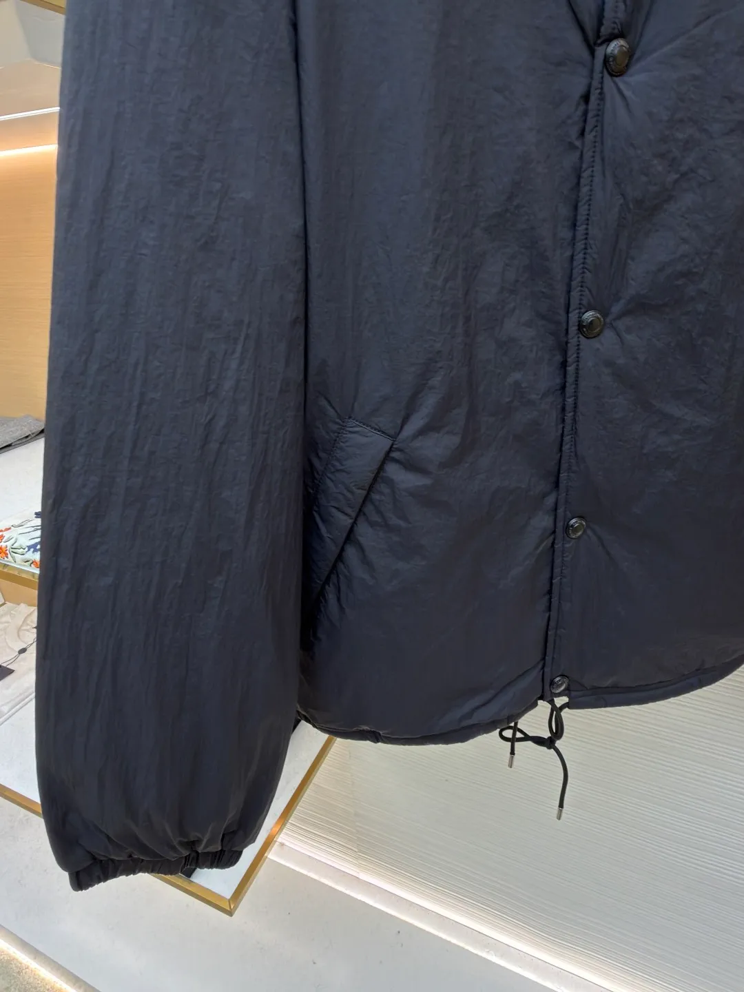 Ralph Lauren red label light cotton jacket (coach jacket) ●Original version: purchased and developed for 2699 ●Item number: 1681910-B90 ●Fabric: high-density nylon taffeta fabric ●Technology: embroidery production ●Color: black ●Version: fitted version ●Weight: about 340 grams ●Size: S-M-L-XL ●Description: The density of high-density nylon taffeta is about 210T-300T, with a smooth and slightly matte surface, excellent windproof and waterproof properties (with DWR water-repellent treatment), strong wrinkle resistance and easy care, in line with the positioning of American casual "durable and practical". Classic dark black, using vat dyeing process (dye penetrates into the fiber), the color fastness reaches ISO level 4 or above, preventing fading or color transfer after washing. The lightweight cotton-polyester blend lining (65% Polyester + 35% Cotton) reduces the friction between the fabric and inner clothing, improving the smoothness of wearing; the blended material combines breathability and durability to avoid the stuffy feeling of pure nylon lining. The classic Coach Jacket has a loose fit with slightly dropped shoulder lines (shoulder width is 1-2cm wider than the standard fit). The length reaches the hips and the sleeves are slightly tapered straight sleeves, taking into account both freedom of movement and casual style. Polo horse logo on the left chest: flat satin stitch embroidery (satin embroidery), embroidery thread: 100% polyester high color fastness embroidery thread (the color number is consistent with the brand's standard red color card), stitch density 12-15 stitches/cm, the outline of the horse logo is clear, and the details of the rider and horse are burr-free. Embroidery positioning: precise positioning through the template (12cm from the neckline and 8cm from the placket) to ensure left and right symmetry and centering. Topstitching technology: The seams of the body are made of double-needle flat seams (needle pitch 3cm/10 needles), the stitches are smooth and straight, the stitching color is dark black (the same color as the fabric), and the stitch width is 0.5cm, which enhances firmness while forming simple decorative lines. The splicing of the front placket and the body: the placket is made of double-layer folding + flat seam splicing, with a thickness of about 0.3cm to ensure that the buttons are not easily deformed when fixed; the edges of the placket are cut with a hot knife to avoid burrs. The sleeves are spliced ​​to the body: the armholes are spliced ​​with round-head keyhole machine seams + flat seams. After splicing, the armholes have a natural curvature and no sense of tightness during movement; the height of the sleeves is about 8-10cm, which is in line with the loose characteristics of casual jackets. Main button: black urea resin button, matte surface treatment, buttonholes are hand-made keyholes (6 strands of cotton thread, keyhole density 8 stitches/mm), and the edges of the eyelets are smooth and without burrs. Collar buttons: The first button on the collar is a hidden button (diameter 12mm), which prevents the collar from opening and improves the neatness of wearing.